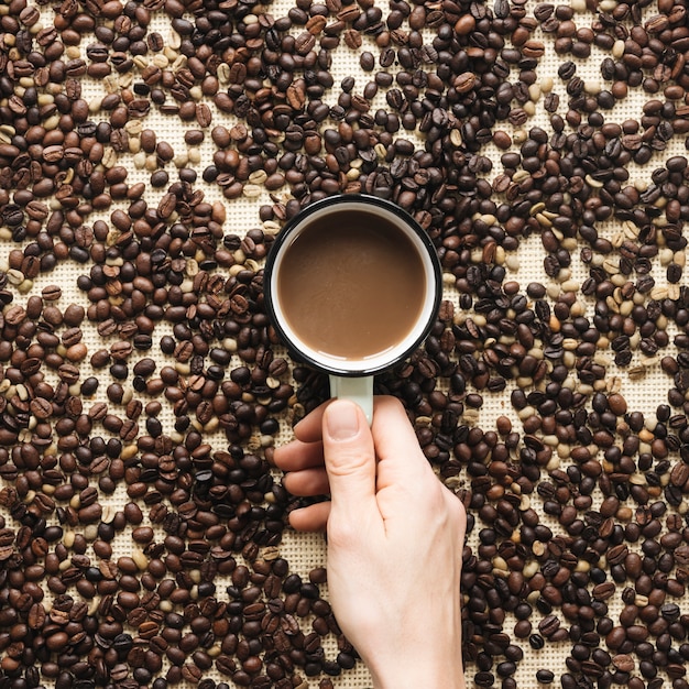 Primer plano de la mano de una persona sosteniendo una taza de café rodeado de granos de café
