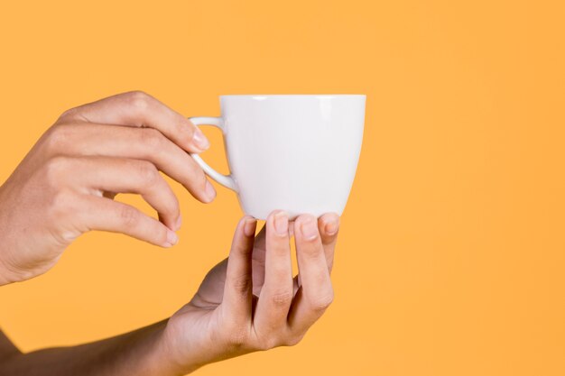 Primer plano de la mano de la persona sosteniendo la taza de té contra el telón de fondo amarillo