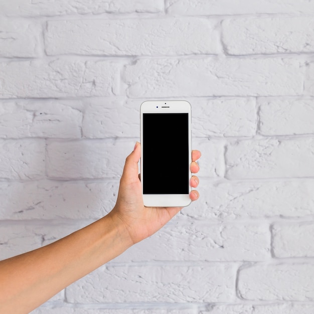 Foto gratuita primer plano de la mano que sostiene el teléfono móvil con pantalla en blanco delante de la pared blanca