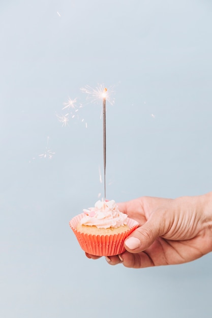 Primer plano de mano sosteniendo cupcake de cumpleaños con bengala sobre fondo gris