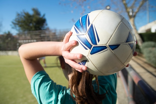 Primer plano de las manos de las adolescentes con pelota. Vista posterior de una joven con una pelota blanca y azul sobre su cabeza en el estadio de la escuela, lista para tirársela a su amiga. Fútbol femenino, concepto de estilo de vida saludable