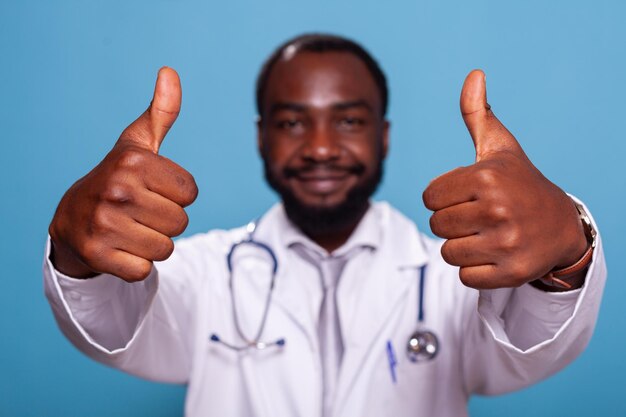 Primer plano de manos dando pulgares hacia arriba de alegre médico afroamericano en uniforme médico con estetoscopio. Detalle recortado de un médico optimista que muestra un gesto de mano para una actitud positiva.