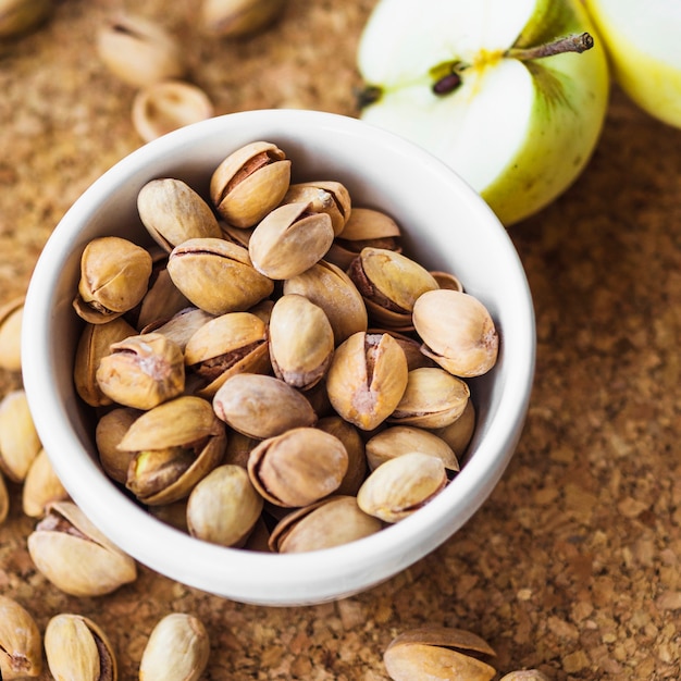 Primer plano de manzana a la mitad con tazón de pistacho en tablero de corcho