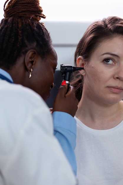 Primer plano de un médico otólogo afroamericano que controla el oído del paciente de la mujer con un otoscopio médico que descubre una infección de otología durante el examen clínico. Terapeuta que trabaja en la oficina del hospital