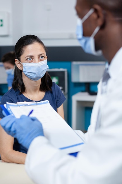 Primer plano de un médico pediatra afroamericano que escribe un diagnóstico enfermo en el portapapeles que explica el tratamiento médico a los padres durante la cita clínica en la oficina del hospital. Terapeuta con mascarilla