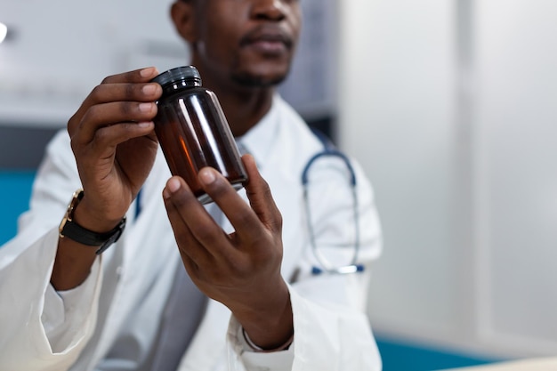 Primer plano de un médico pediatra afroamericano sosteniendo la botella de píldoras en la mano durante la cita clínica en el consultorio del hospital. Hombre terapeuta discutiendo el tratamiento con medicamentos. Servicios de salud