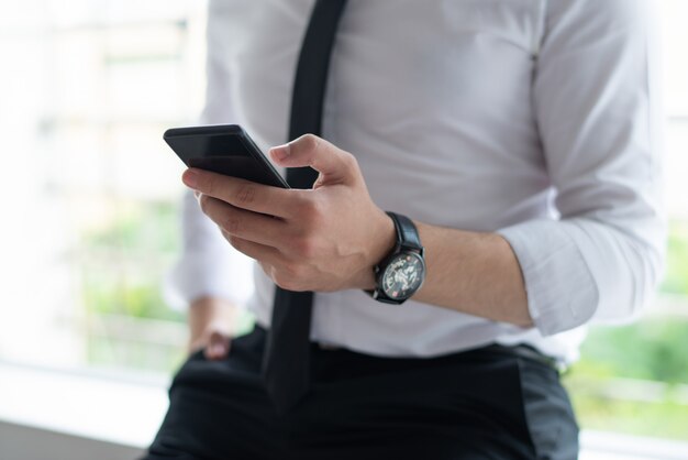 Primer plano de los mensajes de texto del hombre de negocios en el teléfono inteligente y apoyado en el alféizar