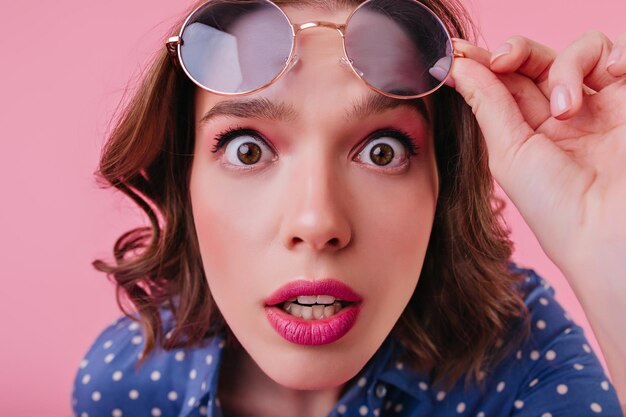 Primer plano de una modelo femenina sorprendida de cabello oscuro sosteniendo sus gafas de sol Retrato de una chica blanca sorprendida aislada sobre fondo rosa con la boca abierta