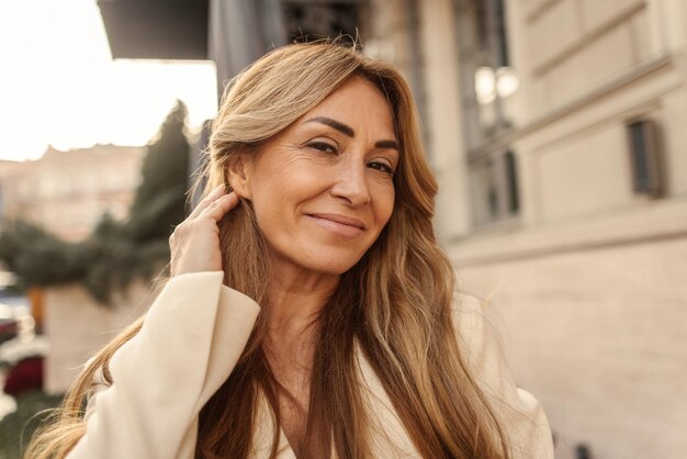 Primer plano de una mujer adulta de piel clara sonriente mirando a la cámara mientras se arregla el cabello rubio detrás de la oreja Personas emociones sinceras concepto de estilo de vida