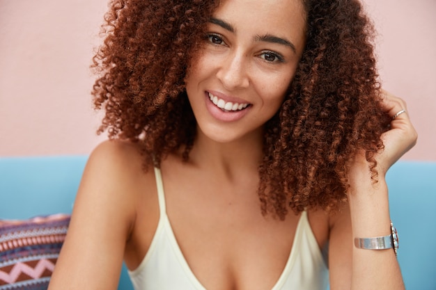 Foto gratuita primer plano de una mujer afroamericana de aspecto agradable que tiene un aspecto encantado, una piel oscura y saludable, posa en el interior del sofá, tiene una expresión feliz