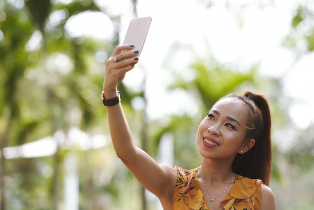 Primer plano de mujer asiática elegante feliz con cola de caballo y maquillaje tomando selfie con smartphone