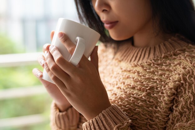 Primer plano de mujer asiática seria en suéter tomando café caliente