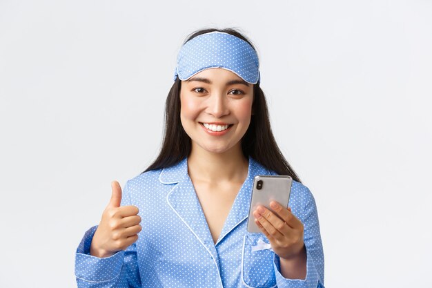 Primer plano de mujer asiática sonriente alegre en pijama azul y antifaz para dormir rastrea su sueño con la aplicación de teléfono inteligente, mostrando el pulgar hacia arriba como usando el teléfono móvil y sonriendo a la cámara complacido.