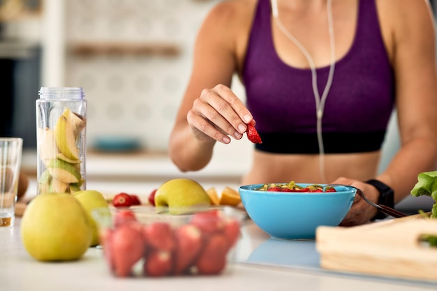 Primer plano de mujer atlética agregando fresas mientras hace ensalada de frutas en la cocina