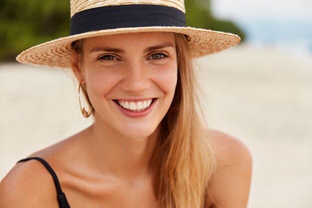Primer plano de mujer atractiva tiene ojos cálidos, amplia sonrisa con dientes blancos y uniformes, usa sombrero de playa, recrea en lujoso resort. Concepto de turismo y viajes de verano. Mujer en isla tropical