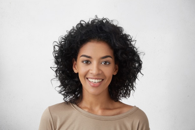 Primer plano de una mujer bonita con dientes perfectos y piel oscura y limpia descansando en el interior, sonriendo felizmente después de recibir buenas noticias positivas.