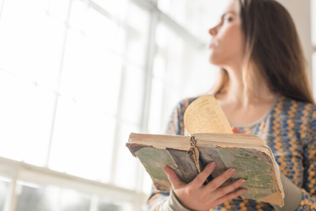 Primer plano de mujer borrosa de pie cerca de la ventana con libro vintage en mano