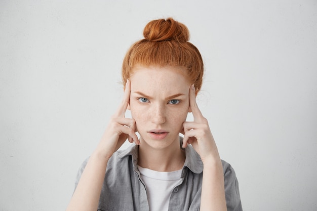 Primer plano de una mujer caucásica joven infeliz estresada que lleva su cabello pelirrojo en un moño, manteniendo los dedos en las sienes, que sufre de dolor de cabeza severo, migraña o se esfuerza por recordar algo