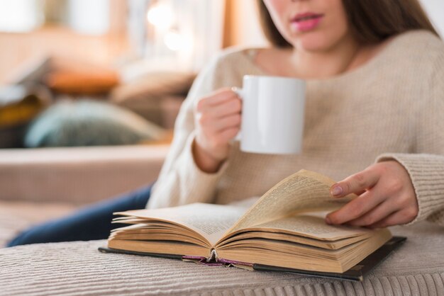 Primer plano de una mujer dando vuelta la página del libro que sostiene la taza de café en la mano