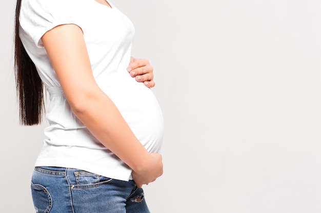 Primer plano de una mujer embarazada irreconocible con las manos sobre la barriga en la pared blanca