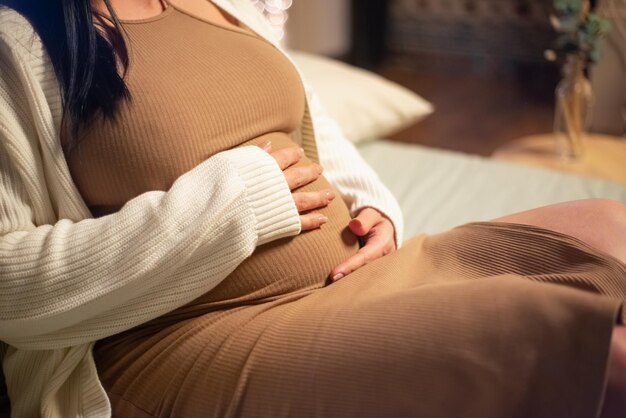 Primer plano de una mujer embarazada sentada en la cama. Mujer con vestido y cárdigan tocando la barriga grande. Embarazo, concepto de expectativa
