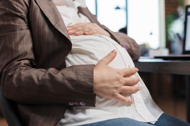 Primer plano de una mujer embarazada tocando la protuberancia del bebé mientras está en el lugar de trabajo de la oficina de la empresa. Futura joven madre sentada en el lugar de trabajo de la empresa de marketing mientras sostiene el vientre del bebé.