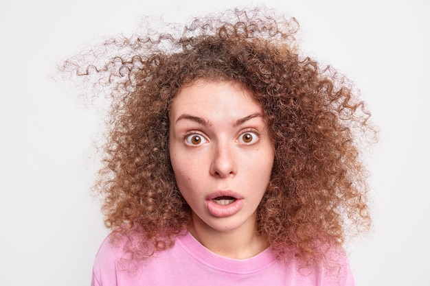 Primer plano de una mujer europea joven y rizada que mira fascinada con los ojos saltones que ha sorprendido que la expresión mantiene la mandíbula caída vestida con ropa casual aislada sobre una pared blanca. Concepto de Omg