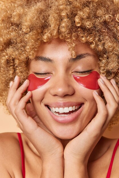 Primer plano de mujer feliz de cabello rizado aplica parches de hidrogel rojo debajo de los ojos para el tratamiento de la piel toca la cara suavemente sonríe ampliamente muestra los dientes blancos se encuentra muy feliz concepto de belleza interior