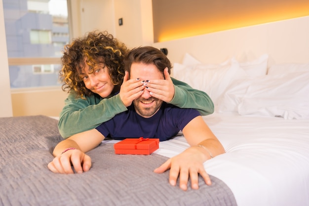 Primer plano de una mujer feliz sorprendiendo a su novio con un regalo en un dormitorio