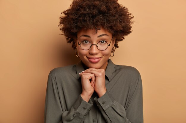 Primer plano de una mujer guapa que tiene una piel sana, mantiene las manos debajo de la barbilla, mira con calma a la cámara, usa gafas redondas y camisa