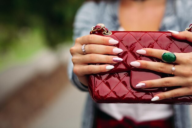 Primer plano de mujer de incógnito con elegante manicura usando joyas sosteniendo una bolsa roja bastante pequeña.