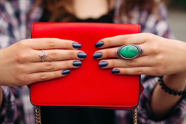 Primer plano de una mujer de incógnito con elegantes uñas oscuras en los dedos cuidados con anillos que llevan una bolsa cruzada compacta roja de color rojo brillante.