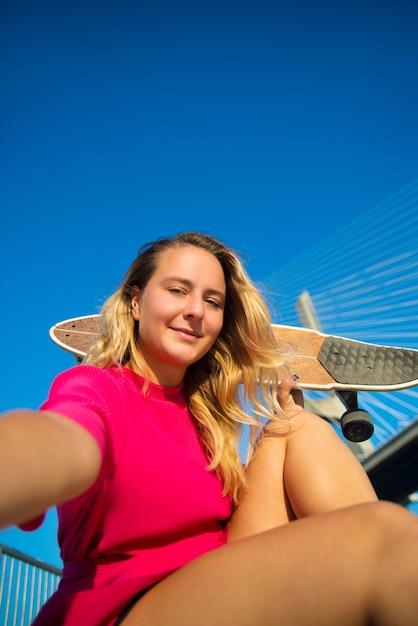 Primer plano de mujer joven atractiva tomando selfie. Hermosa niña sonriente en vestido carmesí con patineta sentada cerca del puente, tomando fotos de sí misma.