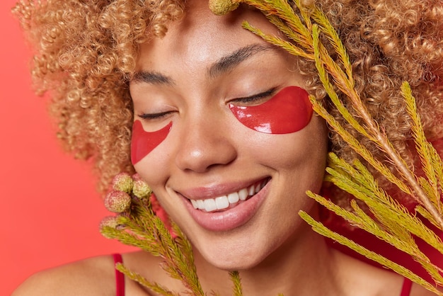 Foto gratuita primer plano de mujer joven complacida con piel sana mantiene los ojos cerrados aplica parches de hidrogel rojo para mimar e hidratar sostiene plantas exóticas cerca de la cara posa interior concepto de belleza