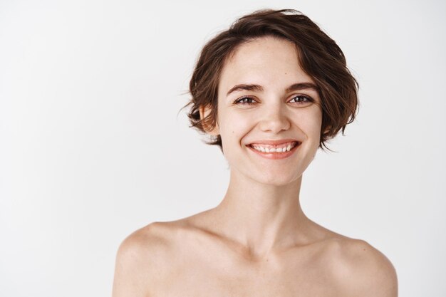 Primer plano de una mujer joven feliz con piel pálida y sin maquillaje, de pie semidesnuda en la pared blanca, sonriendo y mirando feliz. Concepto de cuidado de la piel y belleza femenina