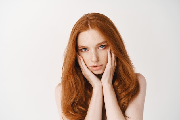 Primer plano de una mujer joven natural con cabello pelirrojo, tocando la cara pálida limpia sin maquillaje y mirando al frente, de pie desnuda sobre una pared blanca