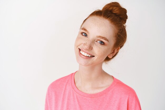 Primer plano de mujer joven pelirroja con pecas y ojos azules tocando limpio, sin piel de maquillaje y sonriendo, de pie en camiseta rosa contra la pared blanca