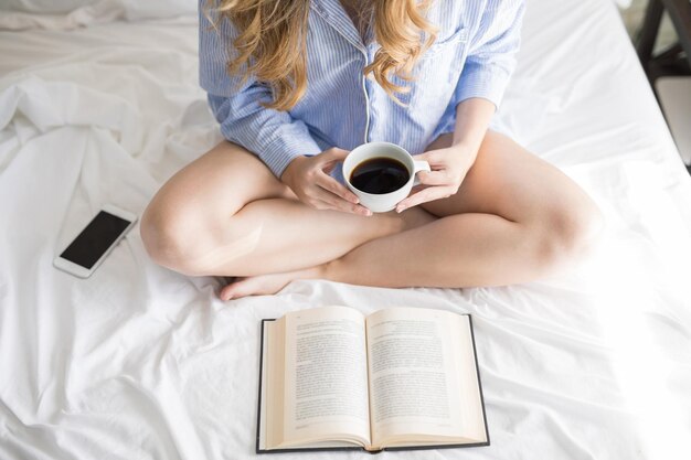 Primer plano de una mujer joven en pijama disfrutando de una taza de café y un buen libro en su cama