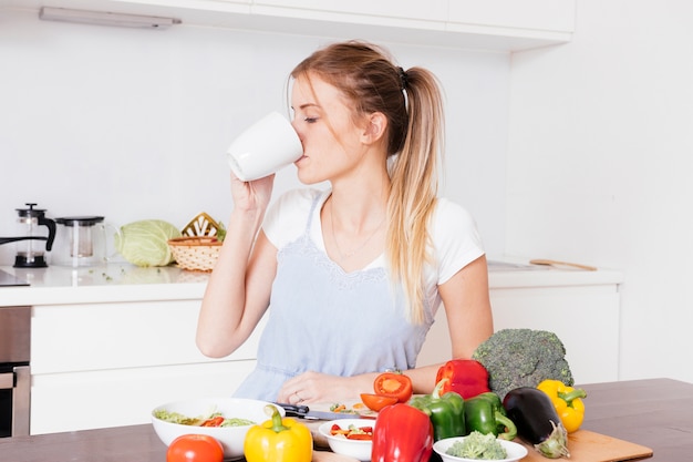 Primer plano de una mujer joven que bebe el café con verduras frescas de colores en mesa de madera