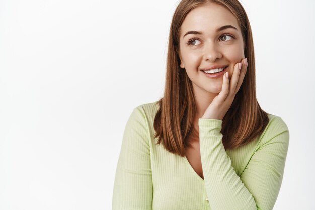 Primer plano de una mujer joven sonriendo, tocando la piel limpia y brillante y mirando el texto de promoción, soñando despierto, concepto de belleza y cuidado de la piel, fondo blanco.