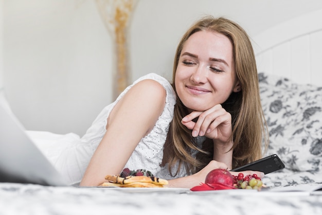 Primer plano de mujer joven sonriente acostado en cama con desayuno mirando portátil