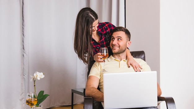 Primer plano de una mujer joven sosteniendo una copa de vino en la mano de pie detrás del hombre usando una computadora portátil