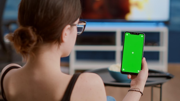 Primer plano de una mujer joven sosteniendo un smartphone vertical con pantalla verde viendo contenido de medios sociales en la sala de estar de casa. Chica usando un teléfono móvil con pantalla táctil con llave croma mirando la pantalla.