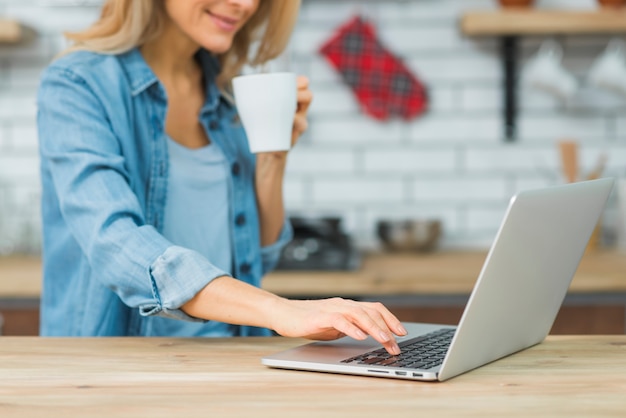 Primer plano de mujer joven sosteniendo una taza de café escribiendo en la computadora portátil sobre la mesa de madera