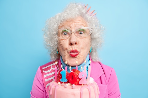 Primer plano de una mujer mayor con cabello gris culry mantiene los labios doblados para soplar velas en el pastel celebra cumpleaños está bien vestido tiene maquillaje brillante disfruta de ocasiones especiales recibe felicitaciones