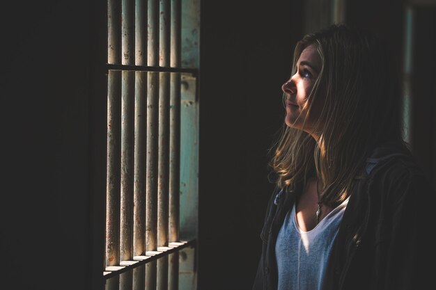 Primer plano de una mujer mirando fuera de una ventana de rejas con el sol brillando en su rostro