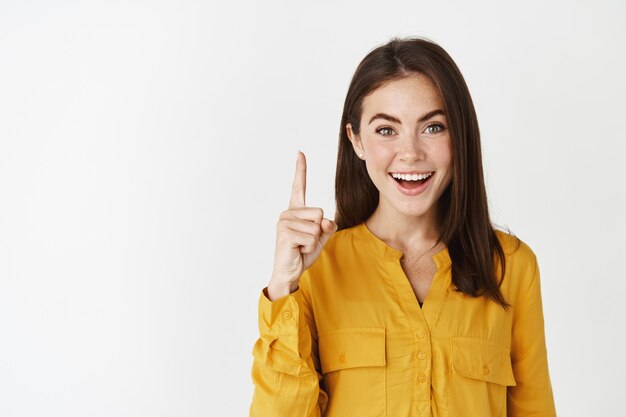 Primer plano de mujer morena divertida y feliz apuntando con el dedo hacia arriba, mostrando publicidad en la parte superior, de pie sobre la pared blanca