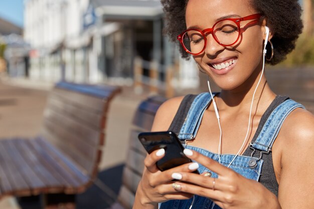 Primer plano de la mujer negra sonriente meloman disfruta de la radio en línea, sostiene el teléfono móvil, está conectado a auriculares digitales, usa gafas ópticas, modelos contra la calle al aire libre borrosa