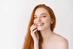 Foto gratuita primer plano de mujer pelirroja feliz con piel pálida perfecta, riendo y mostrando los dientes blancos, de pie desnuda en la pared del estudio