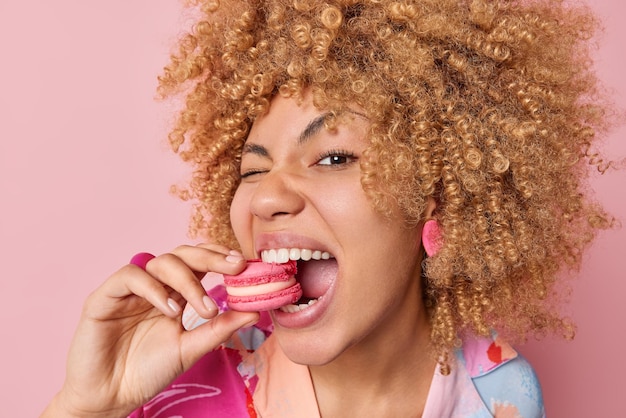 Primer plano de mujer de pelo bastante rizado come deliciosos macarrones franceses disfruta postre panadería dulce comida deliciosa mantiene la boca ampliamente abierta poses contra fondo rosa Concepto de alimentación poco saludable